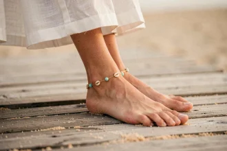 Bracelet cheville en chaîne dorée avec perles turquoise et coquillages sur une cheville féminine posée sur un ponton en bois au bord de la plage