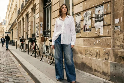 Jeune femme portant un jean large indigo et une chemise oversize blanche dans une rue pavée parisienne, style urbain saison