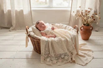 Robe de baptême bohème en dentelle ivoire pour bébé fille, posée dans un panier en osier avec tissu mousseline crème dans un décor champêtre naturel