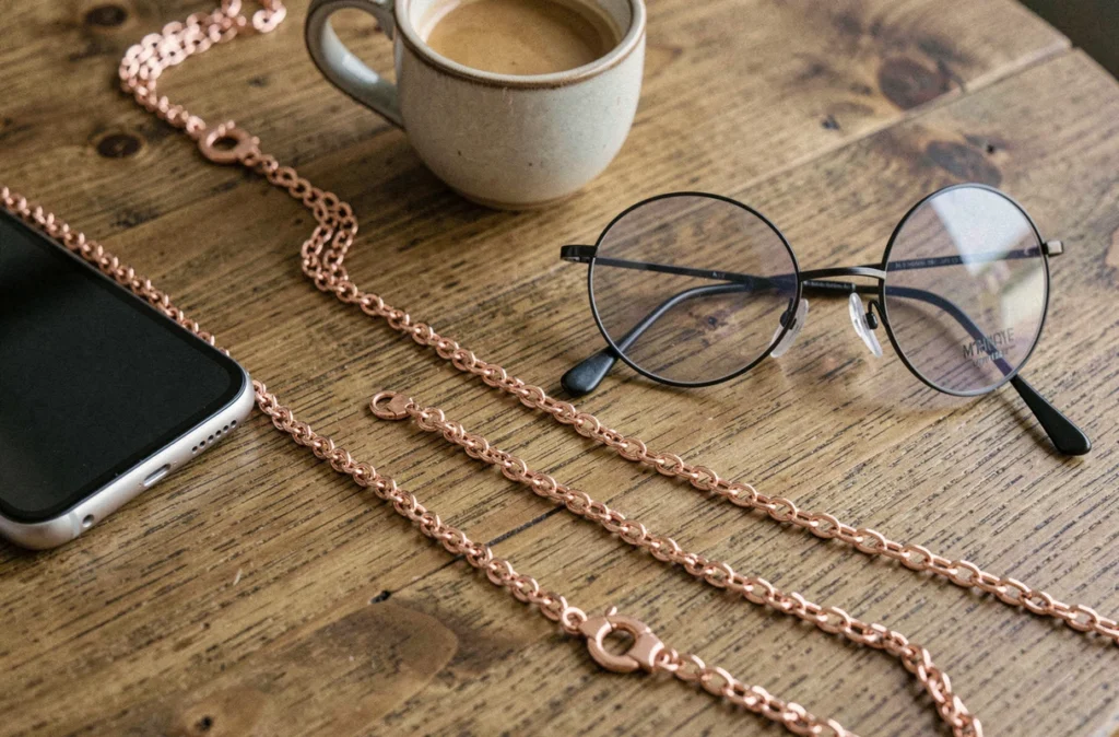 Chaîne porte-téléphone et lunettes en or rose posée sur une table de café avec smartphone et lunettes rondes